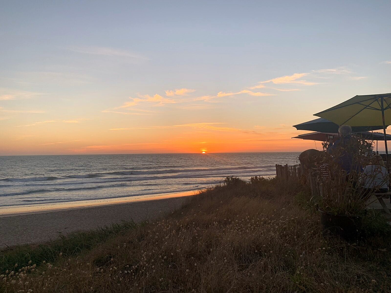 Sonnenuntergang am Atlantikstrand mit Strandbar-Sonnenschirmen und Dünengras im Vordergrund
