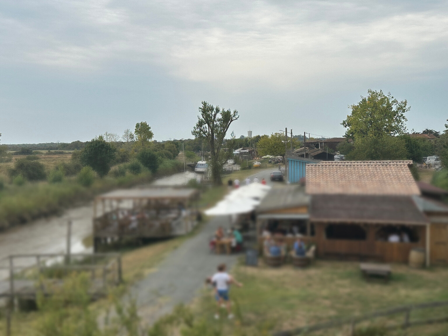 Le Kayak Café am Chenal du Gua in Saint-Vivien-de-Médoc: Holzterrasse über dem Wasser, Sonnenschirme, gefüllte Restaurant-Plätze, Pinienwald-Horizont